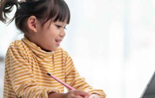 Smiling young girl using a tablet while writing, demonstrating how virtual occupational therapy builds fine motor skills and focus at home.