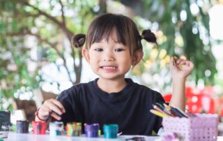 A young girl smiles playfully while painting with bright colors at a table, showing confidence and joy through creative occupational therapy.