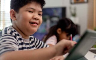 A young boy smiles while using a digital tablet, with another child writing in the background—highlighting focus tools for children with ADHD