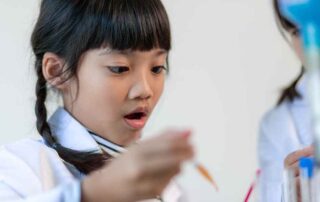 A young girl in a lab coat looks surprised and engaged as she participates in a science-based activity, exploring with focus and curiosity.