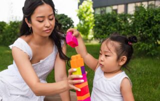 A young toddler girl plays outdoors with her mother, stacking colorful blocks together, supporting coordination and gross motor skills through play.