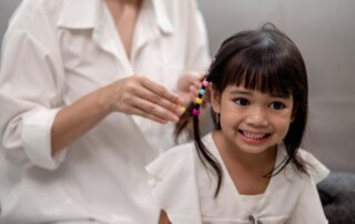 A young girl smiles while her hair is braided with colorful beads, demonstrating fine motor and self-care skill building in a supportive routine.