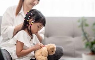 A young girl practices fine motor skills by tying a ribbon on a teddy bear during a home activity, guided by her caregiver.