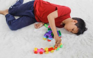 A young boy lies on a soft rug while playing with colorful building blocks and toy vehicles during a focused occupational therapy activity.