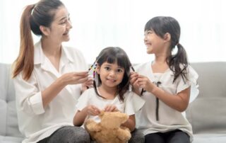 A mother and sister help braid a young girl’s hair with colorful beads, supporting fine motor skills and social bonding through everyday routines.