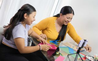 A smiling mother and daughter engage in an origami craft activity together, building fine motor skills and coordination in a joyful setting.