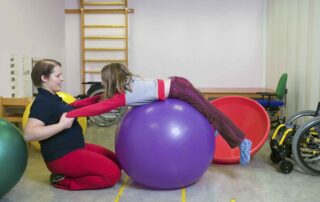 A child with a physical disability doing core-strengthening therapy using a purple exercise ball, assisted by a therapist at OrbRom Center in Phnom Penh