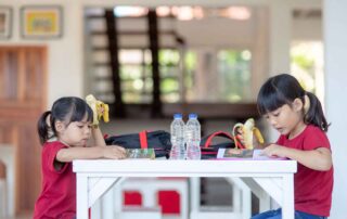 Two young girls in red shirts reading and eating bananas at a white table, representing healthy routines and literacy support in Phnom Penh preschools.